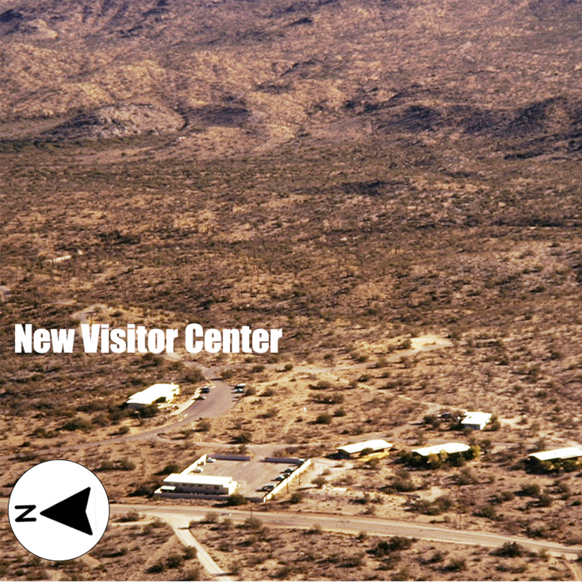 Black & white aerial photo of a historic visitor center pre-60s. A small building sits among a landscape of desert flora. A small direction arrow in the lower left points up for North.