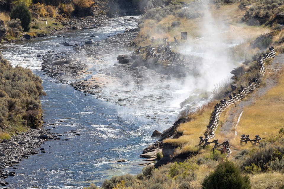 A river winds through the mountains. A large group of people are swimming in it and walking on a paved path beside it.