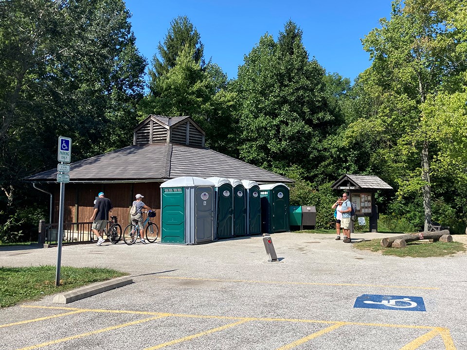Five green portable restrooms lined up in front of a brown wooden building near the edge of a paved parking lot, green trees in the background.