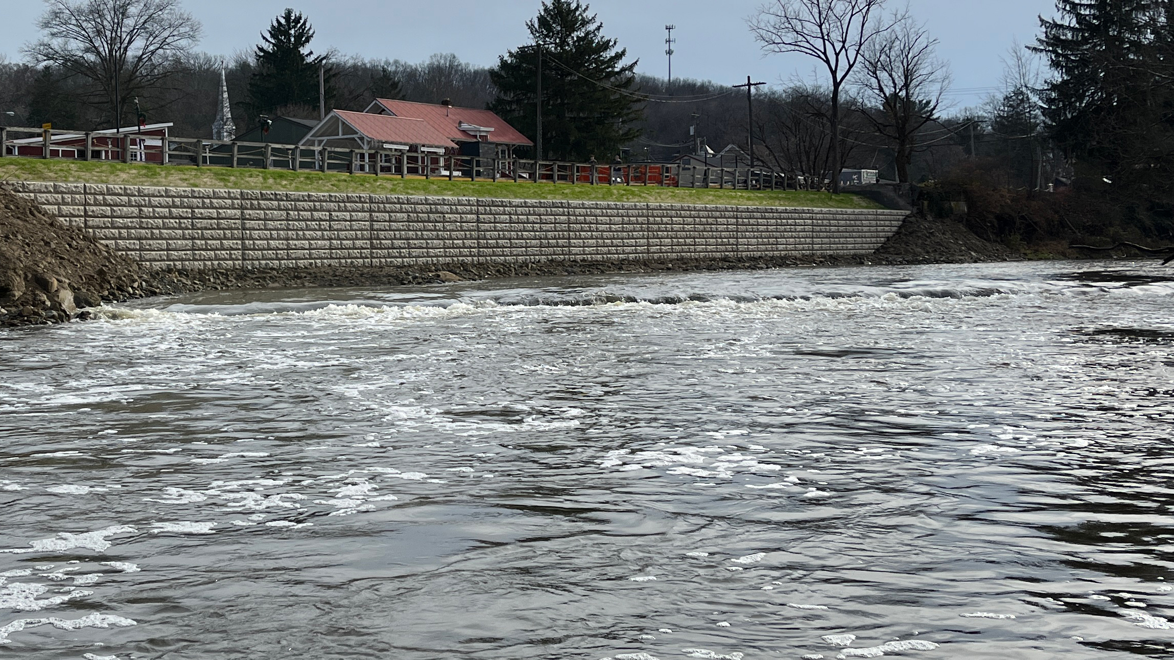 Two people paddle a red raft through riffles on a river; buildings and a wooden fence along the riverbank above.