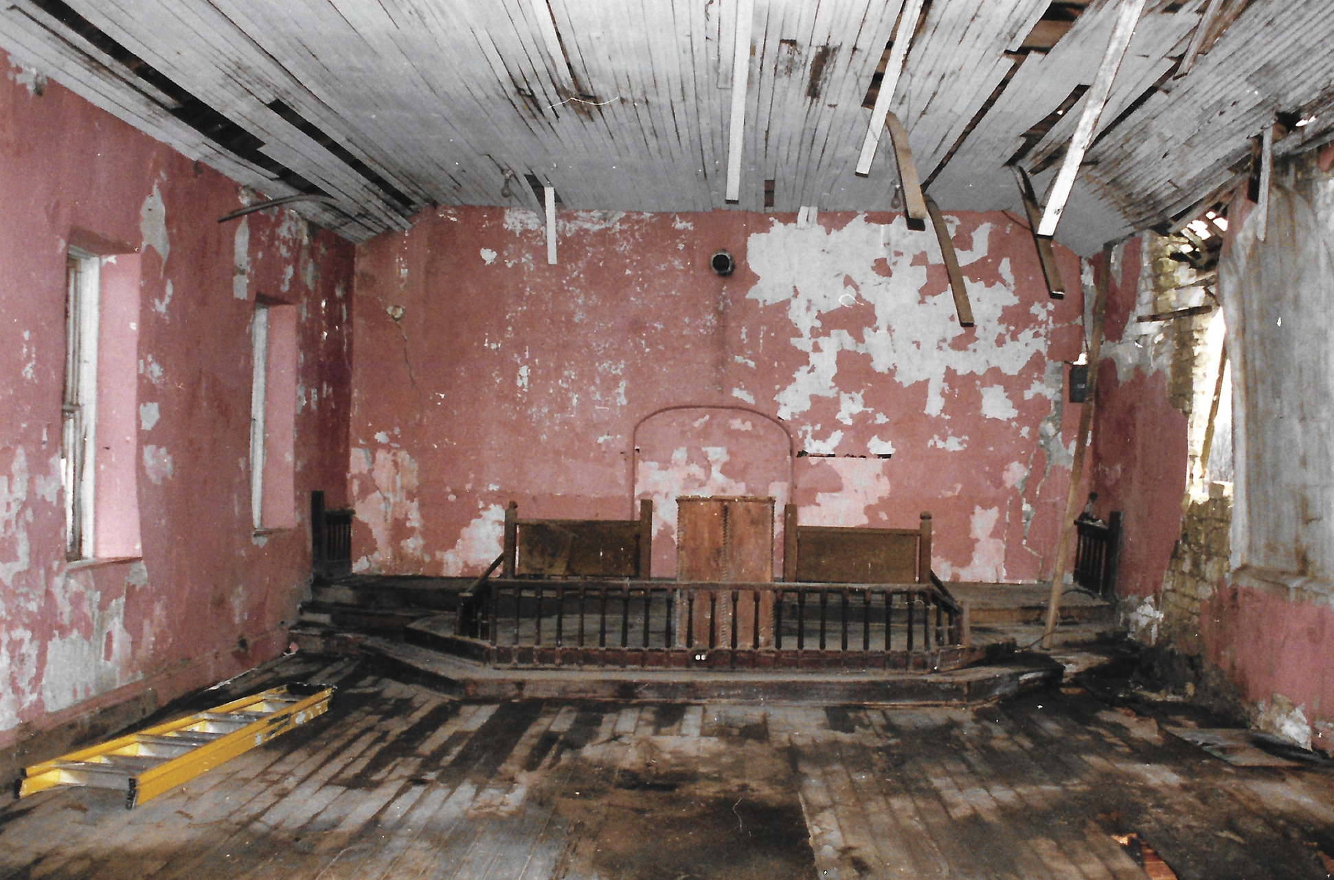 Interior of room with chipped pink walls and a hole in the wall on the right. The wooden floor looks like its rotting and leads to a raised wooden platform with a wooden railing on the front. The ceiling is white thin boards; many boards are dangling.