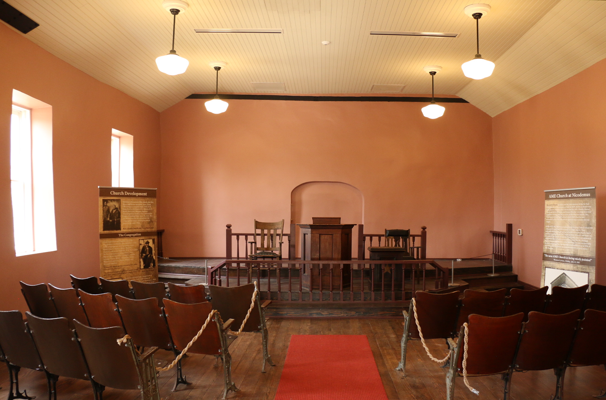 Interior of room with chipped pink walls and a hole in the wall on the right. The wooden floor looks like its rotting and leads to a raised wooden platform with a wooden railing on the front. The ceiling is white thin boards; many boards are dangling.