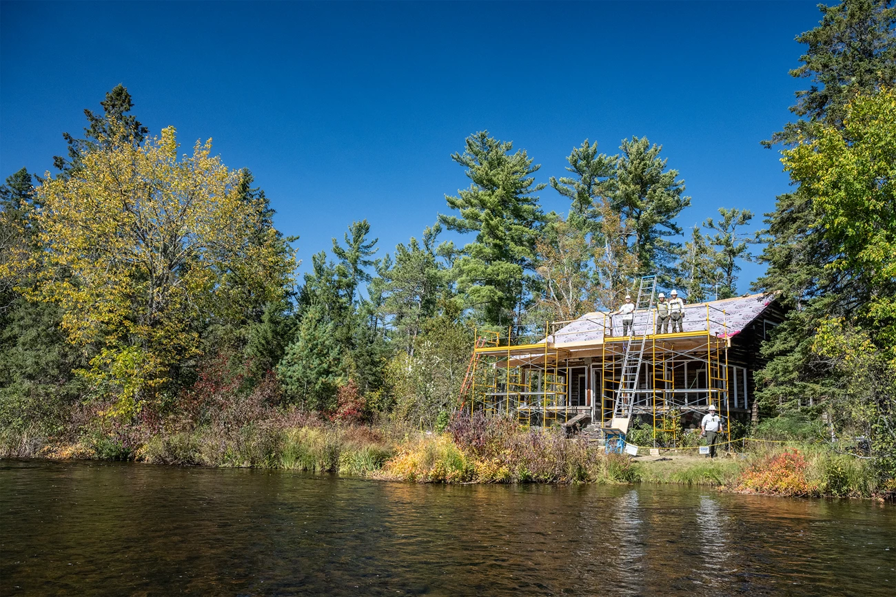 Nestled into a colorful early-autumn treeline along a river is a cabin covered in yellow scaffolding. Construction workers stand on its roof and on the ground near by.