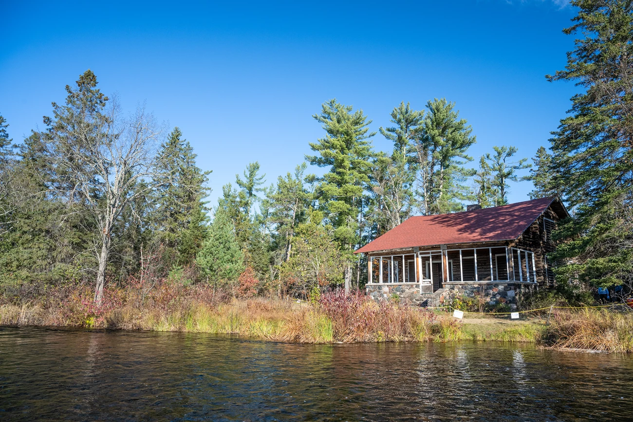 Nestled into a colorful early-autumn treeline along a river is a cabin covered in yellow scaffolding. Construction workers stand on its roof and on the ground near by.