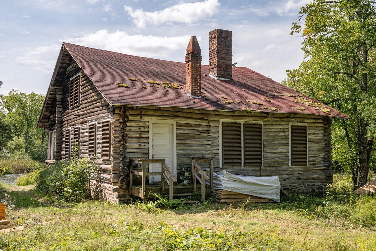An old log cabin with faded and sagging brick-red roof with patches of moss on it. A small porch leads to a white door. Several shuttered windows are on the two visible sides of the cabin.