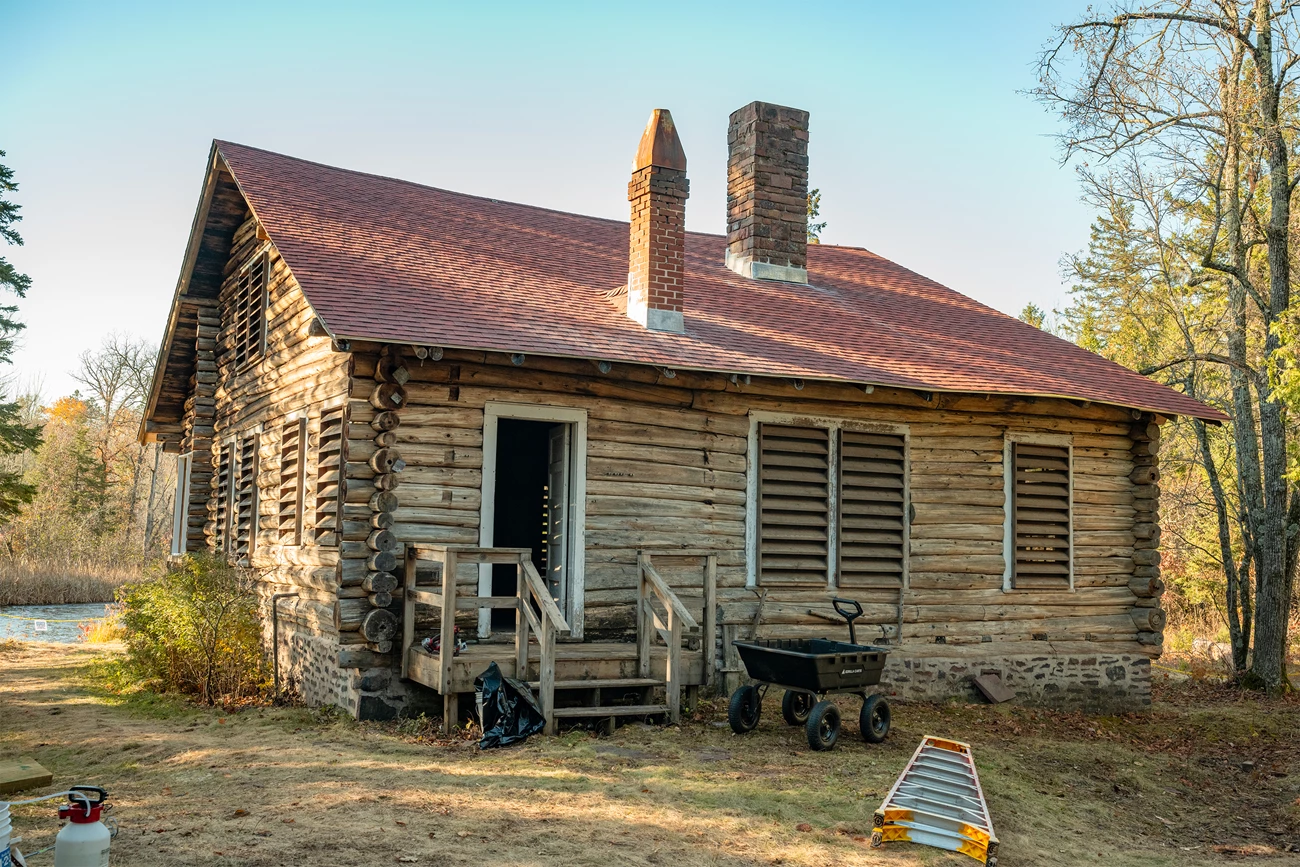 An old log cabin with faded and sagging brick-red roof with patches of moss on it. A small porch leads to a white door. Several shuttered windows are on the two visible sides of the cabin.