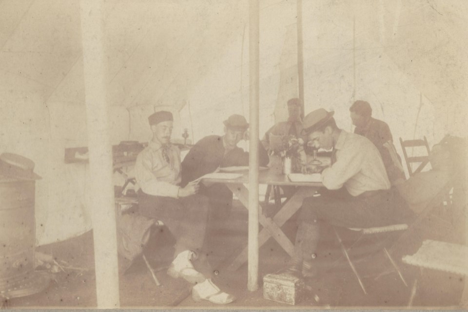 five men sit around a table under canopy tent writing in journals