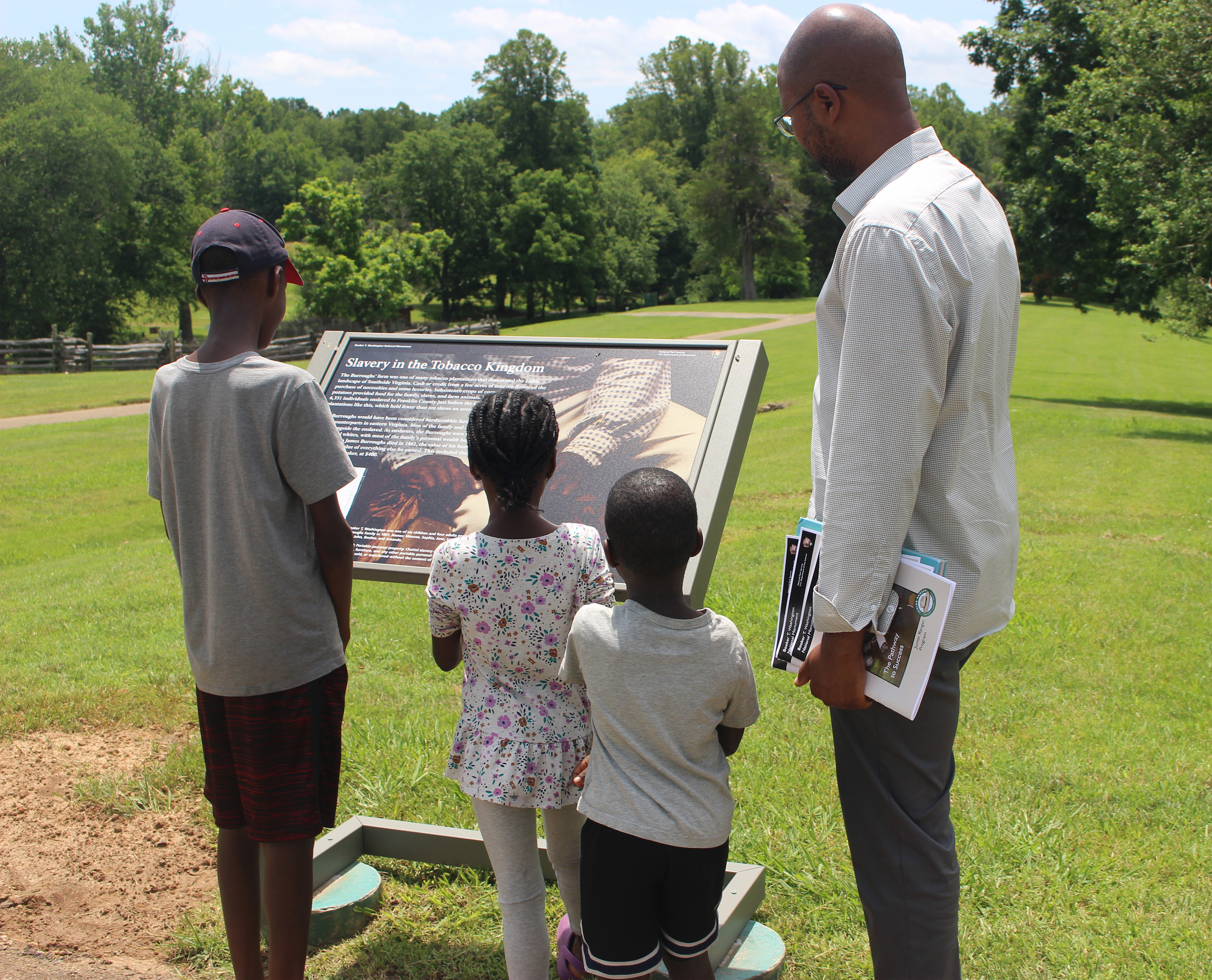 Three children and one adult stand in front of an interpretive wayside depicting slavery.