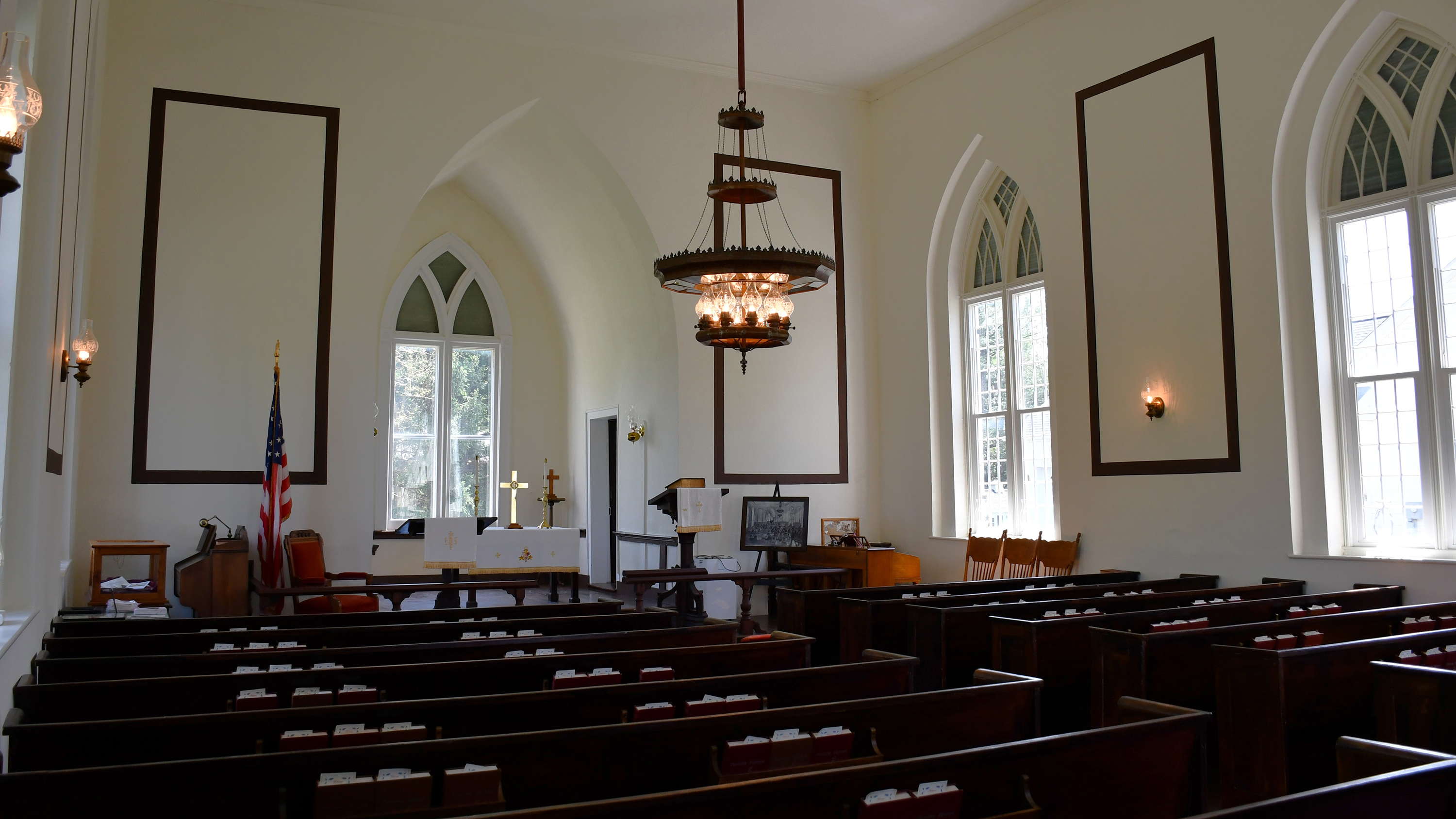 Chapel's interior lined with wooden pews. A chandelier hangs from the ceiling, a large bible rests on a lectern in the altar.