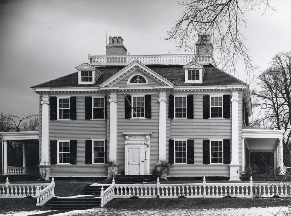Black and white photograph of large mansion with symmetrical facade, two and a half stories, with porches at sides and low railing along front.