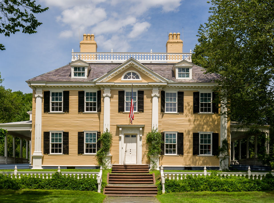 Black and white photograph of large mansion with symmetrical facade, two and a half stories, with porches at sides and low railing along front.