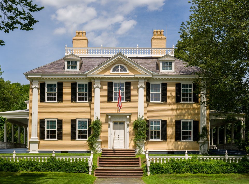 Black and white photograph of large mansion with symmetrical facade, two and a half stories, with porches at sides and low railing along front.