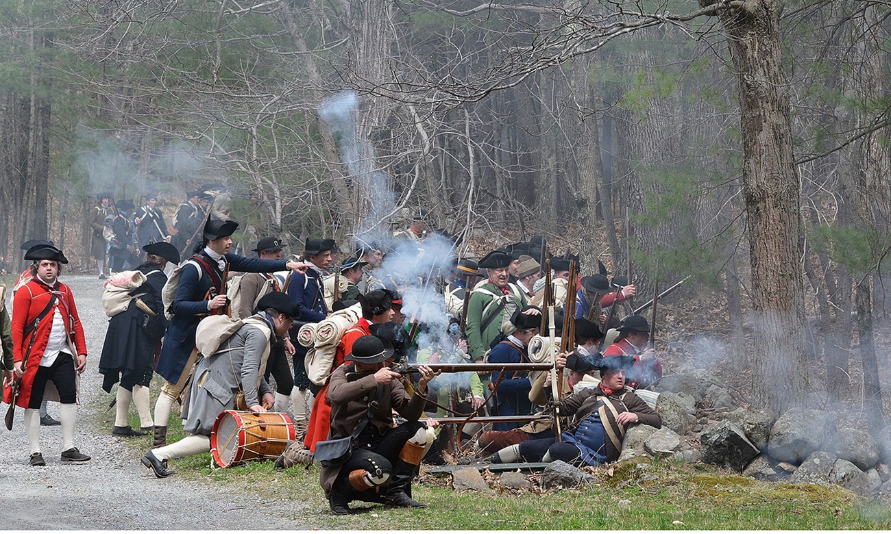 Militia soldiers take cover behind a stone wall and fire their muskets at an unseen enemy