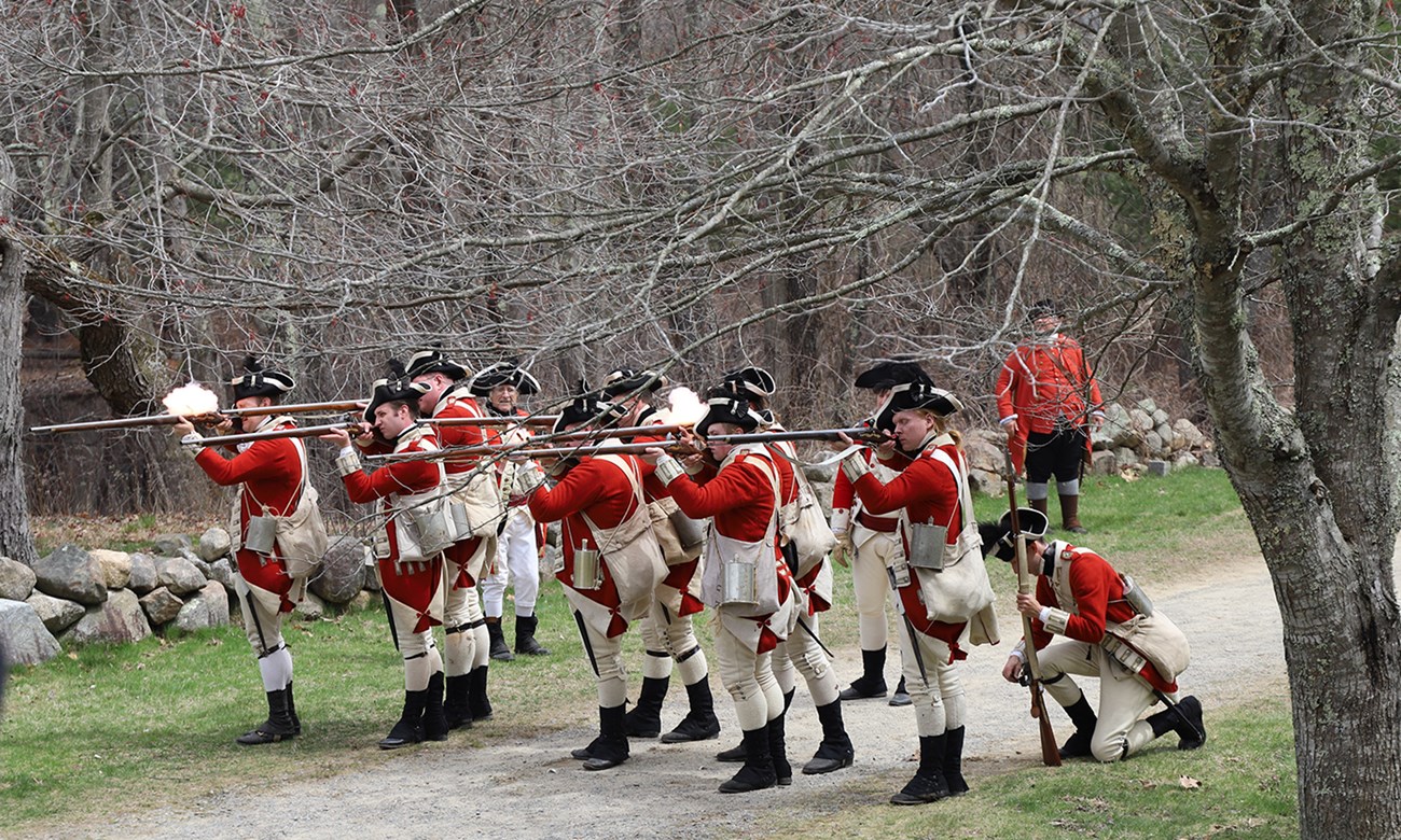 Militia soldiers take cover behind a stone wall and fire their muskets at an unseen enemy