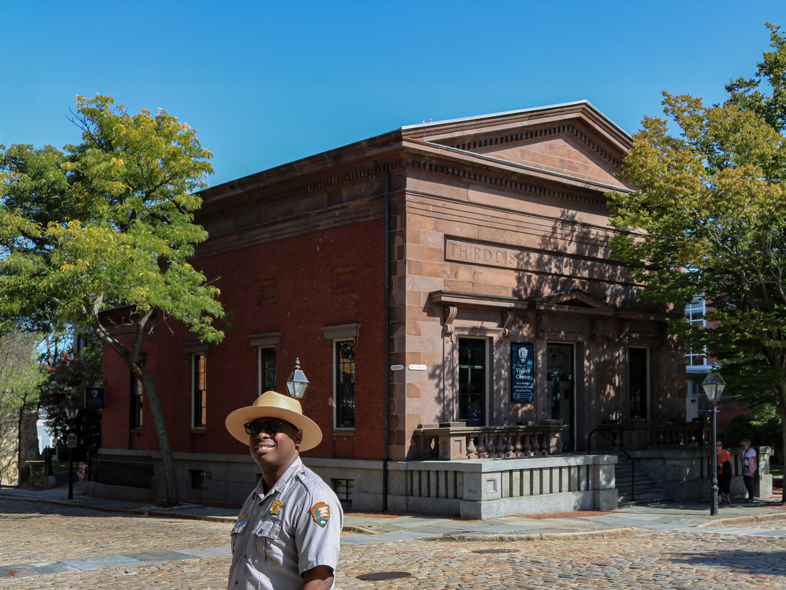 A historic black-and-white photo of the New Bedford Institution for Savings building with stone columns and a pediment. A young man stands in front, and horse-drawn carriages are parked on the cobblestone street.
