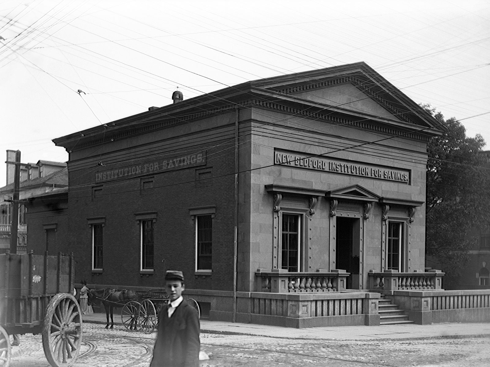 A historic black-and-white photo of the New Bedford Institution for Savings building with stone columns and a pediment. A young man stands in front, and horse-drawn carriages are parked on the cobblestone street.