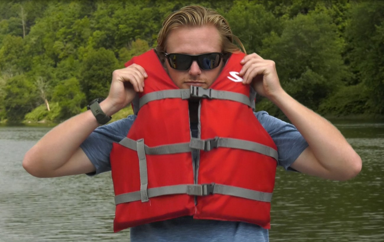 Blond man wearing lifejacket. Lifejacket buckles are closed, but jacket fits very loosely. Man lifts jacket and the jacket's shoulders easily can be lifted over his ears.