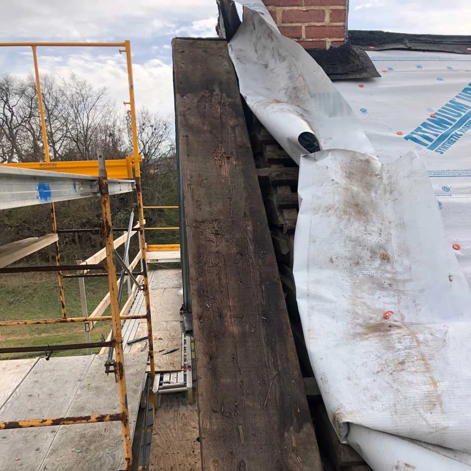 Peak of the end of the roof exposed. The large gable board is dark brown and has a number of places where holes exist and wood is rotten.