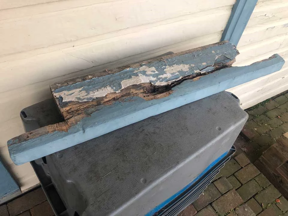 The peeling paint on the windowsill is light blue in color. Where the windowsill matched the wall of the home, there is extensive damage, holes, cracks, and rotted wood.