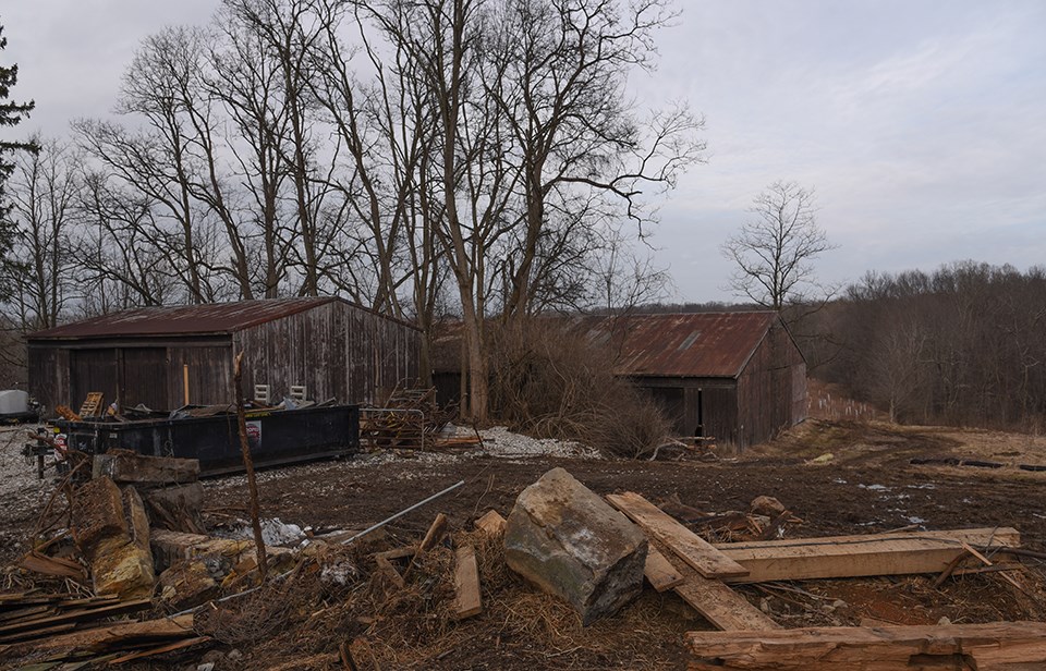 Large sandstone blocks and wooden beams lay scattered by a dumpster, shed, and barn.