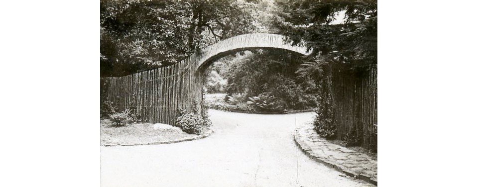 Black and white photo of entry arch spanning a driveway, connected to wooden fencing.