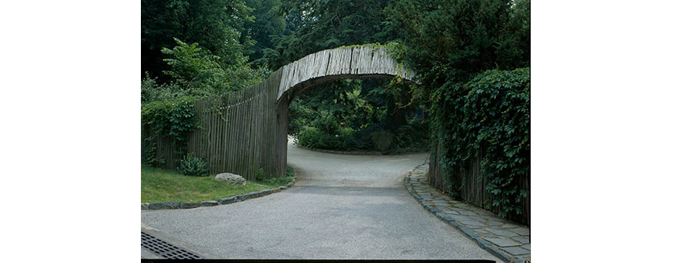 Black and white photo of entry arch spanning a driveway, connected to wooden fencing.