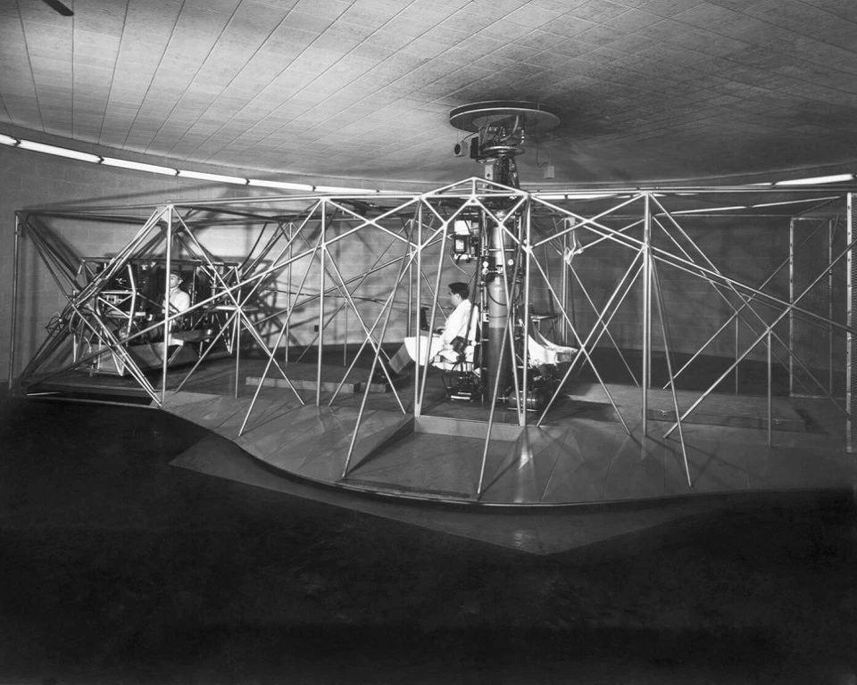 Two people sit back to back at the center of a spinning apparatus composed of several thin beams between two larger horizontal boards