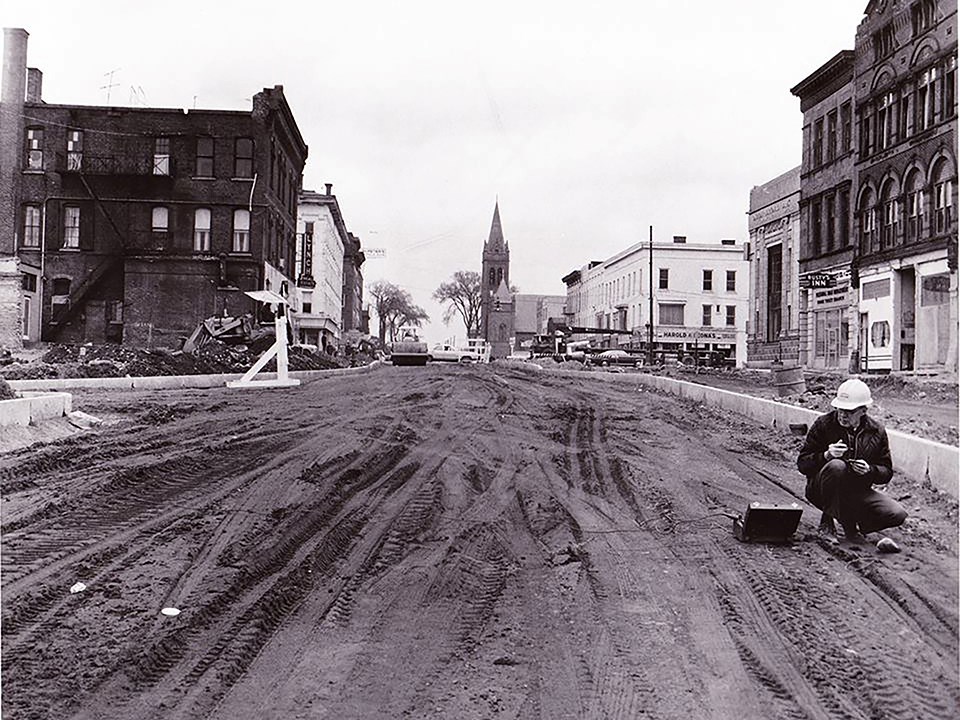 A city street covered in dirt with construction going on along the length. A church steeple in the distance.