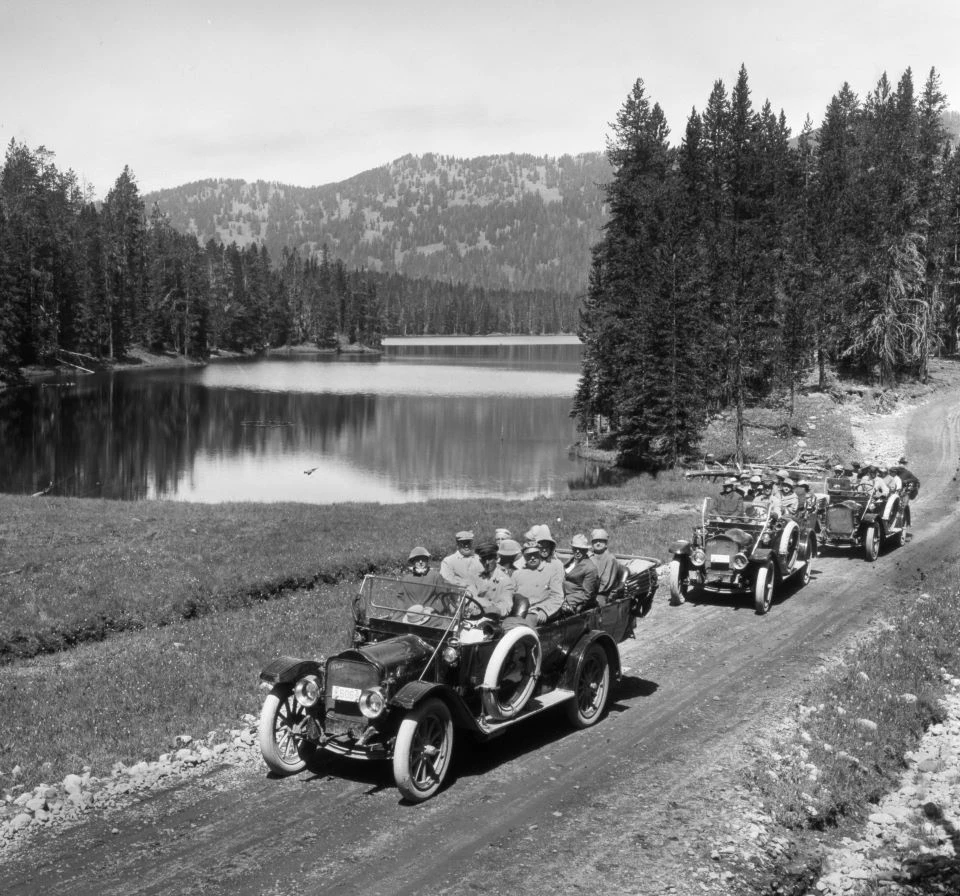 Black and white photo of three open top old-fashioned cars full of people on a dirt road with a lake, trees, and mountains.