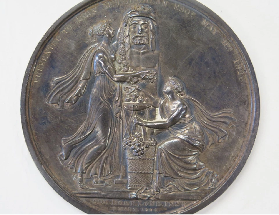 A dull silver medal showing two figures in flowing clothes kneeling on either side of a statue.
