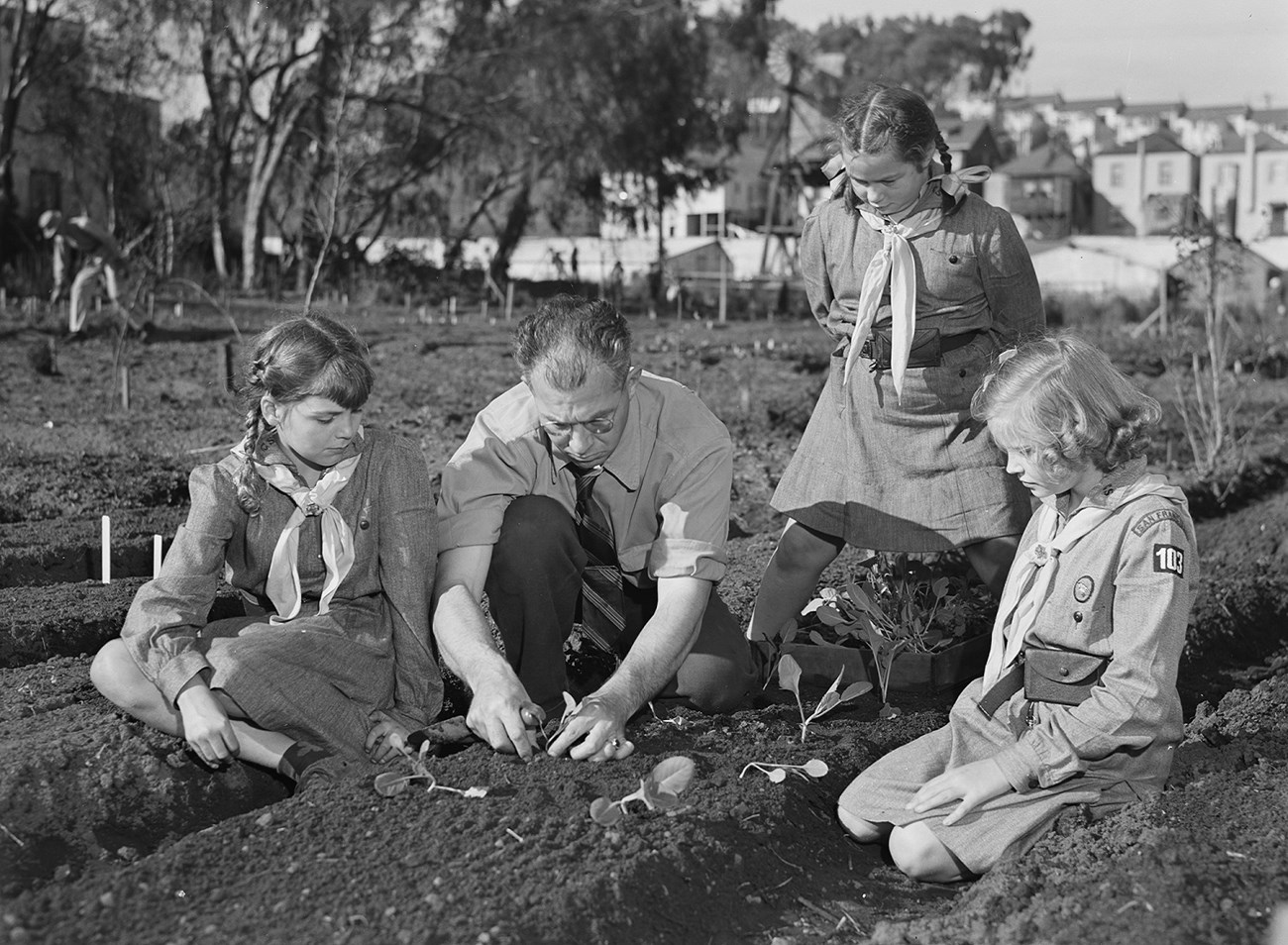 Black and white photo of a white man in a tie kneeling in a garden with plants. Three white girls in Girl Scout uniforms look on.