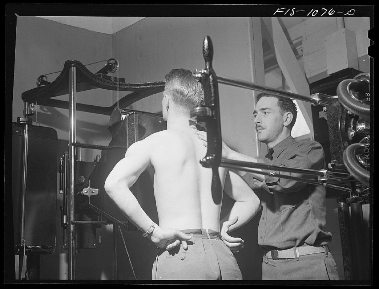Black and white photo. Middle-aged white man with a mustache conducts a physical exam of a shirtless young man who faces away from the camera with his hands resting behind his hips. The examinee is blond and muscular. Metal measuring devices surround them