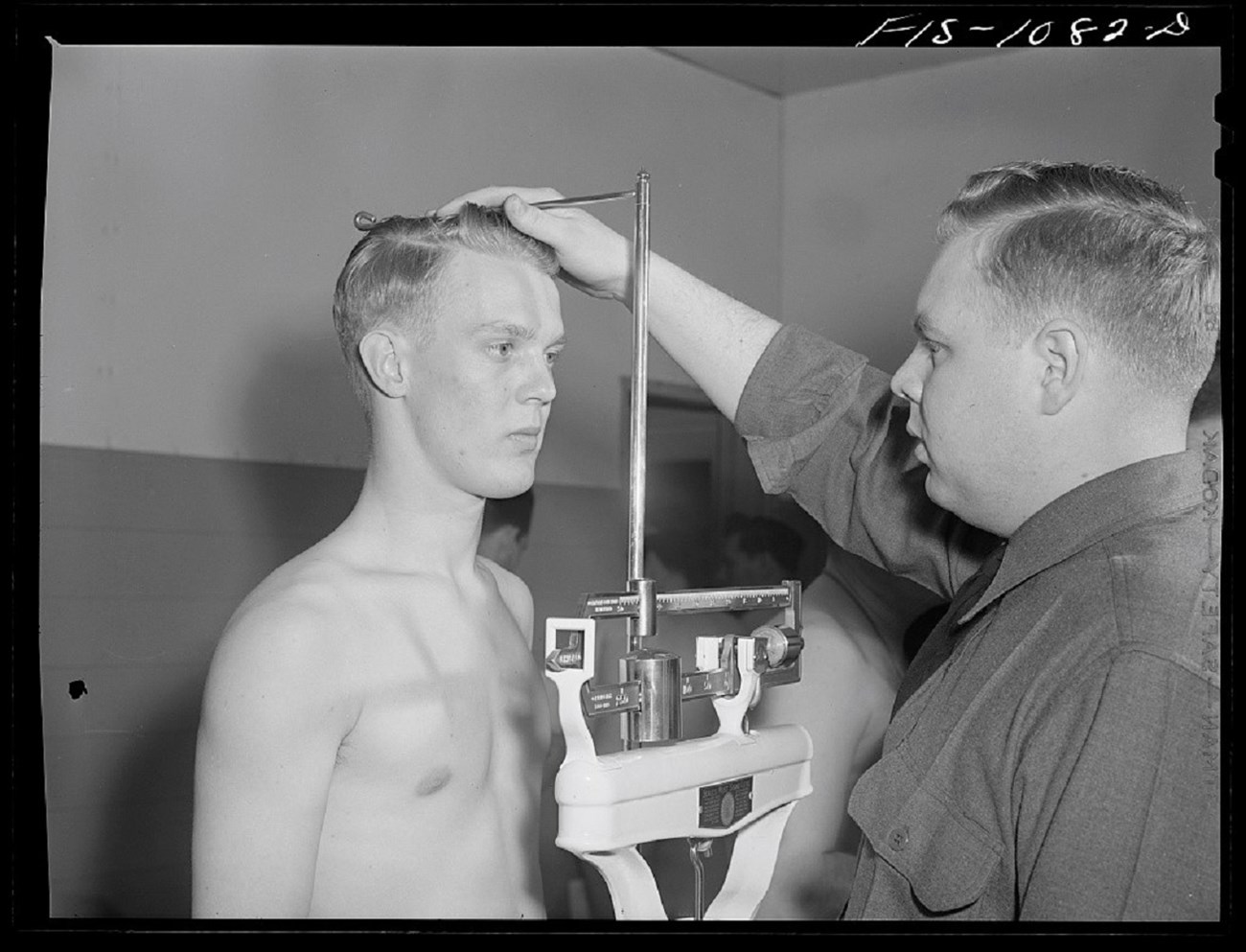 Black and white photo. Middle-aged white man with a mustache conducts a physical exam of a shirtless young man who faces away from the camera with his hands resting behind his hips. The examinee is blond and muscular. Metal measuring devices surround them