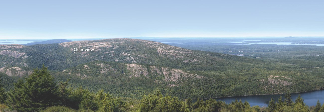 A view from the mountain with pine trees, exposed granite, and a lake visible. The sky is gray and landmarks in the distance grow increasingly obscured the further they are through the hazy air. Text reads: hazy view