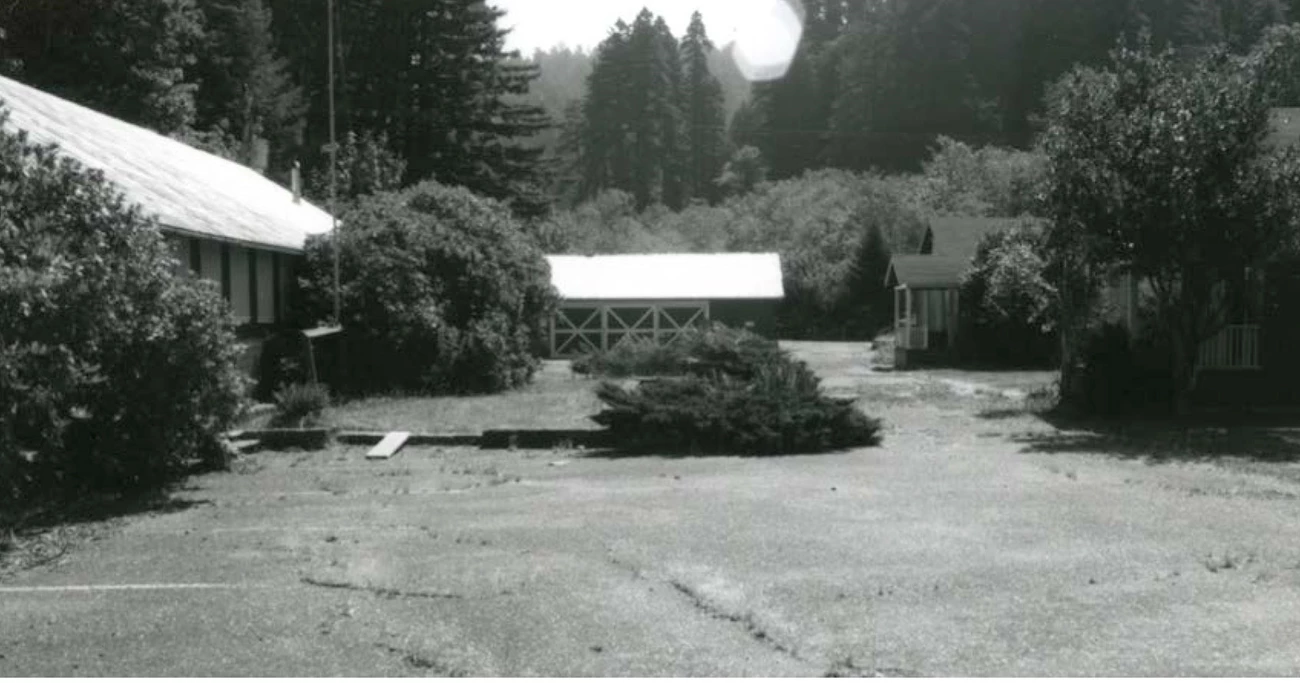 One-story buildings form a perimeter around an area of lawn and asphalt. Some shrubs grow at the foundations and a pond is in the center.