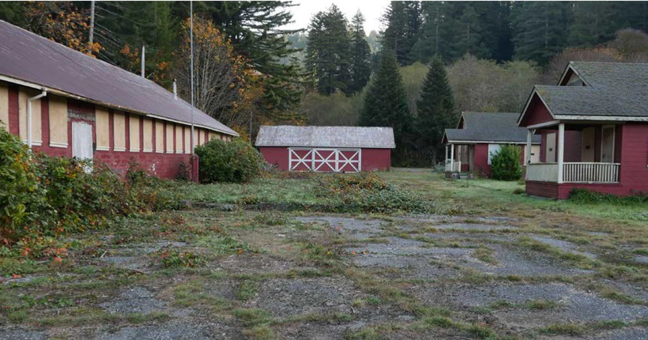 One-story buildings form a perimeter around an area of lawn and asphalt. Some shrubs grow at the foundations and a pond is in the center.
