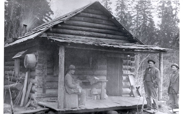three men standing on or near the front porch of a small log cabin home