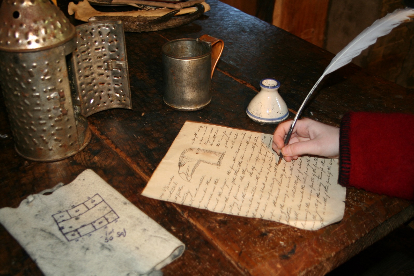 A hand holding a quill pen writes on historic parchment in front of a candle lantern. A hand holding a quill pen writes on historic parchment in front of a candle lantern.