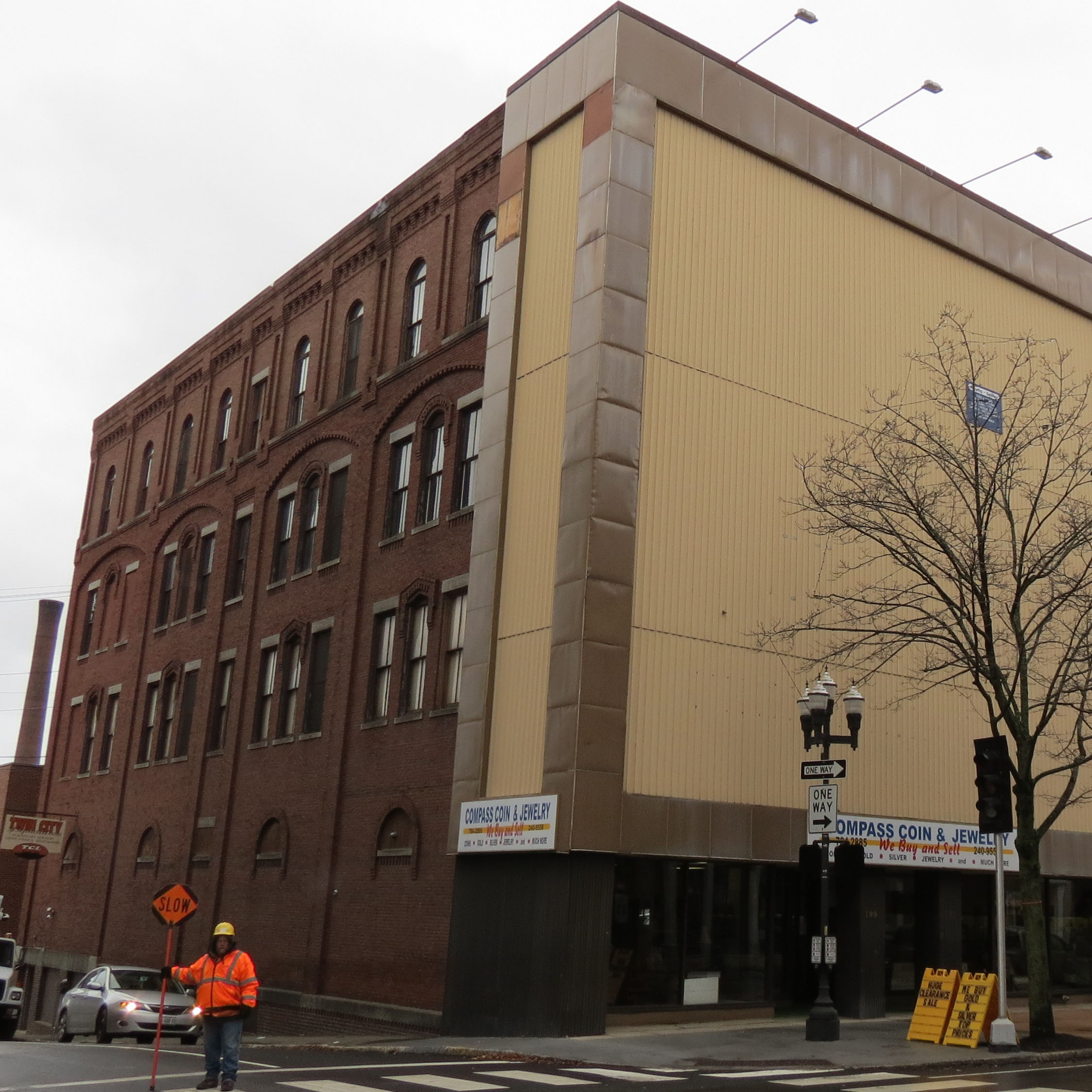A red brick building with multiple windows on a street. A smokestack is in the background, with a blue sky and white clouds.