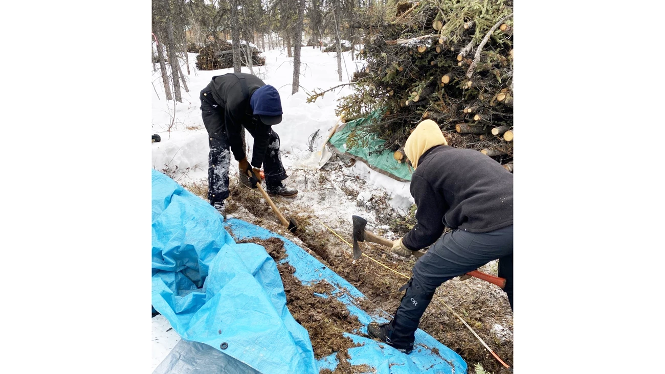 Two people dig a trench in a snowy area, piling soil onto a blue tarp.