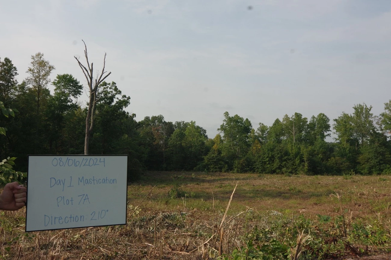 Sign reads 7/16/2024 Plot 7 2024 Pre Mastication Direction 210 in front of a green shrub covered landscape.