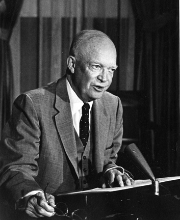 A man in a suit seated behind a desk and microphone A man in a suit seated behind a desk and microphone