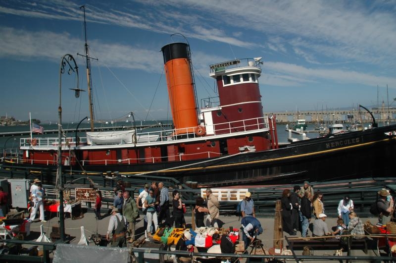 red and black tugboat is tied to pier. A large group of people is waiting to board. 