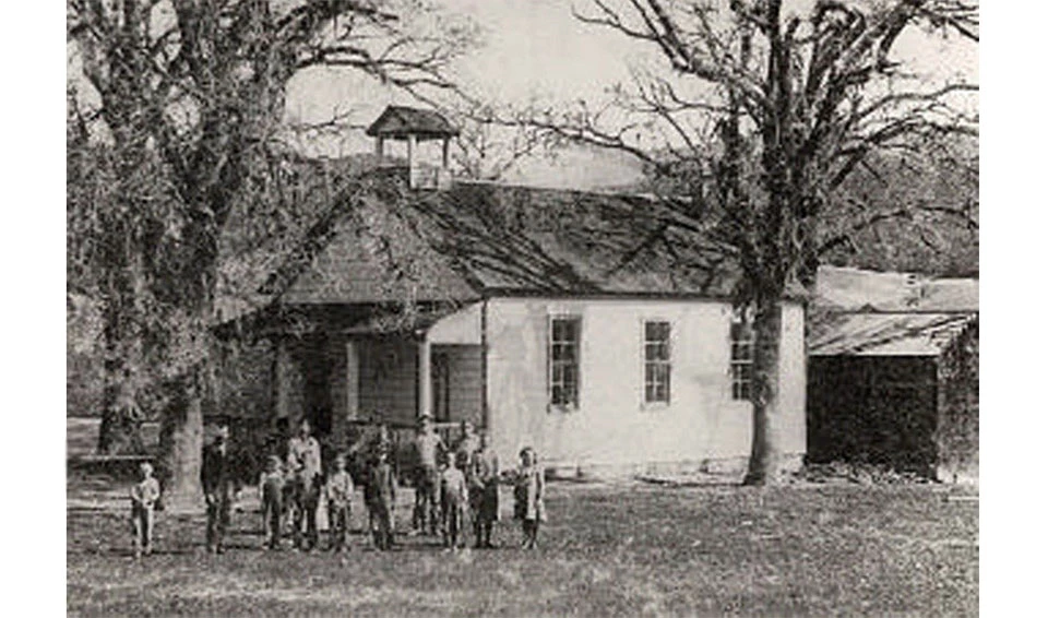A group of students stand in a grassy area near trees in front of a school building with a porch and bell