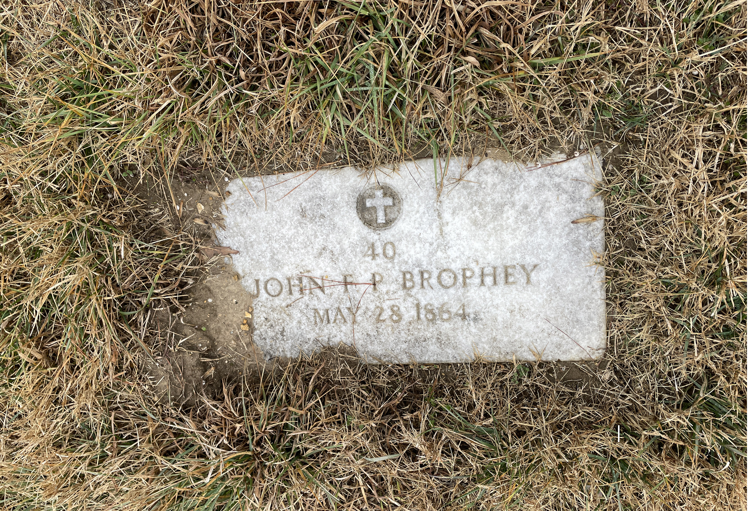 A rectangular headstone with the inscription "John F P Brophey, May 28, 1864" is partly covered by dirt and turf