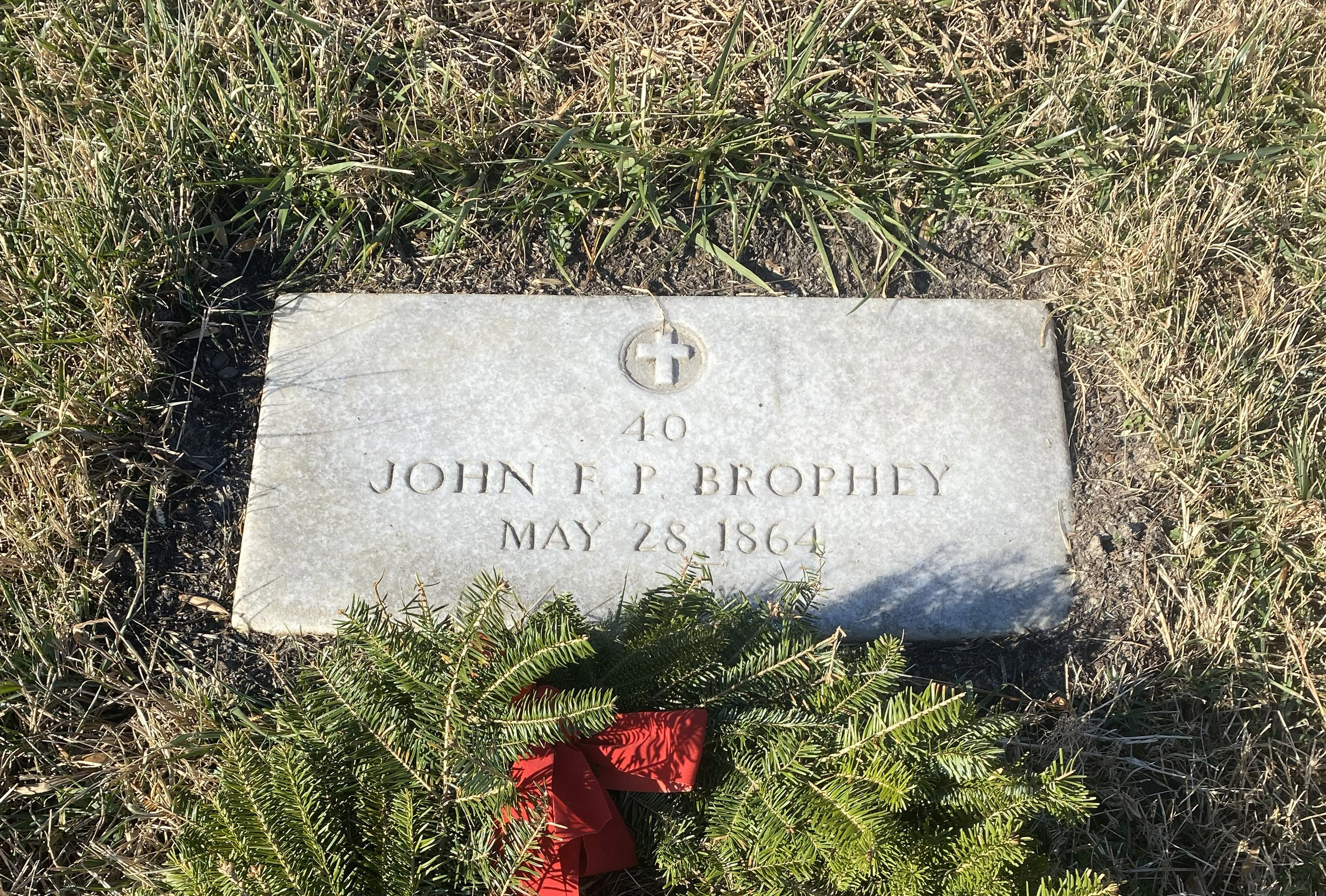 A rectangular headstone with the inscription "John F P Brophey, May 28, 1864" is partly covered by dirt and turf