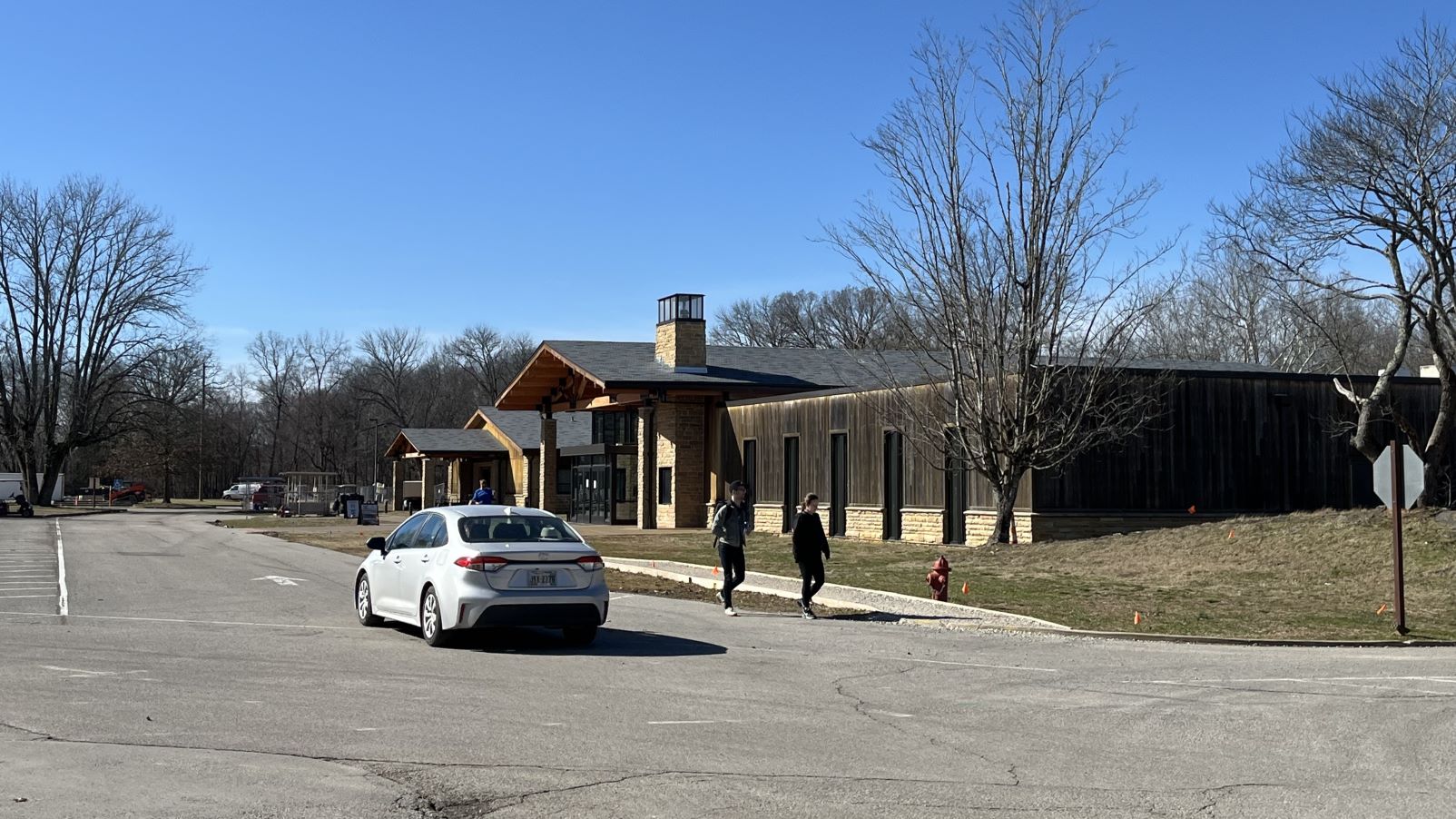 A car drives near pedestrians walking along the side of the road in front of a large wooden framed building.