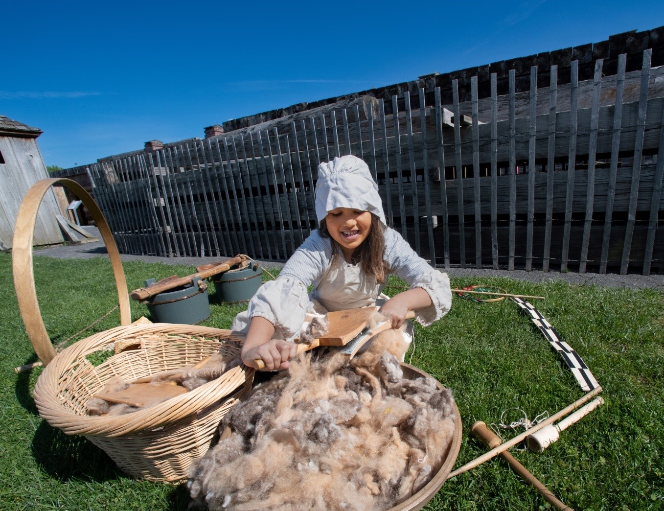 A little girl in historic dress cleans wool in a wooden basket. 