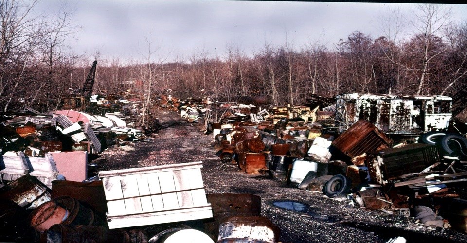 A faded photograph of a dirt road with several puddles and trash stacked on either side including metal containers, horse trailer, damaged furniture and more.
