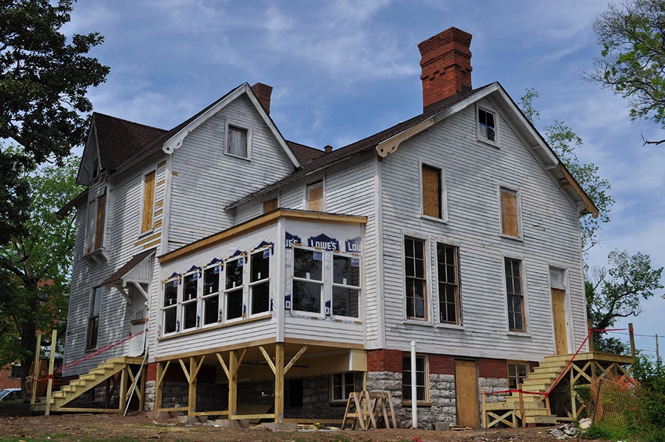 House with white siding, windows boarded up and construction taking place.